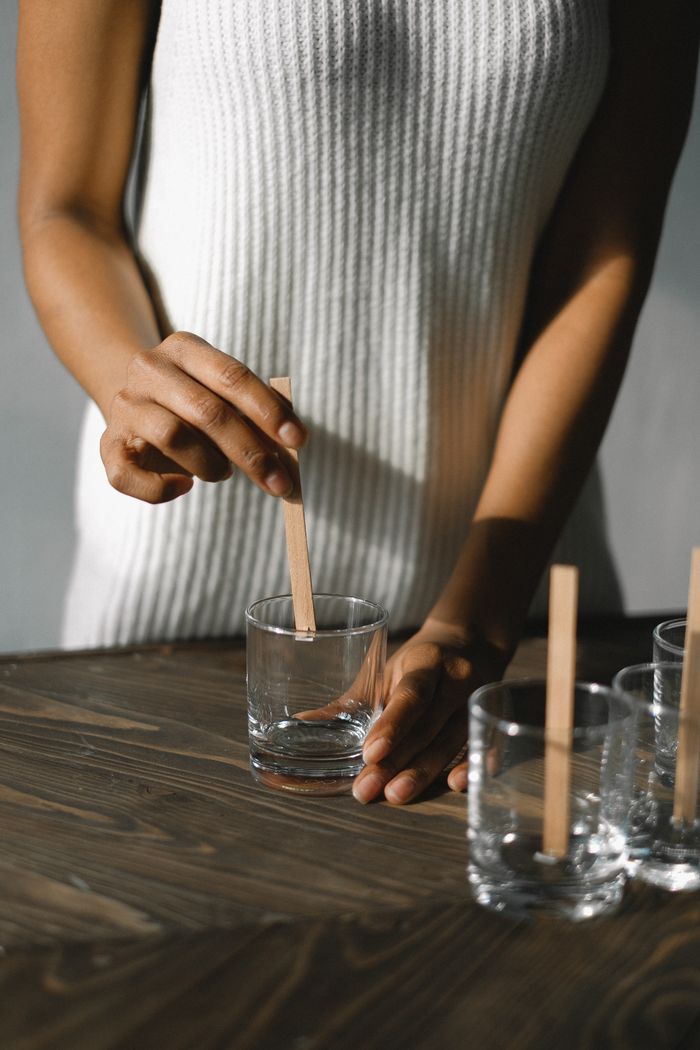 Unrecognizable skilled African American female securing wooden wicks to bottom of glass candle mold while standing at table in light room