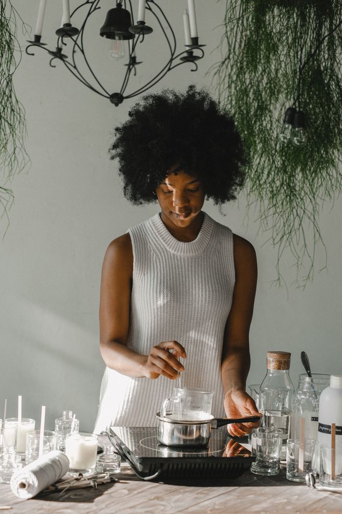 Cheerful African American female artisan heating wax for handmade candles in glass flask on cooker in workshop