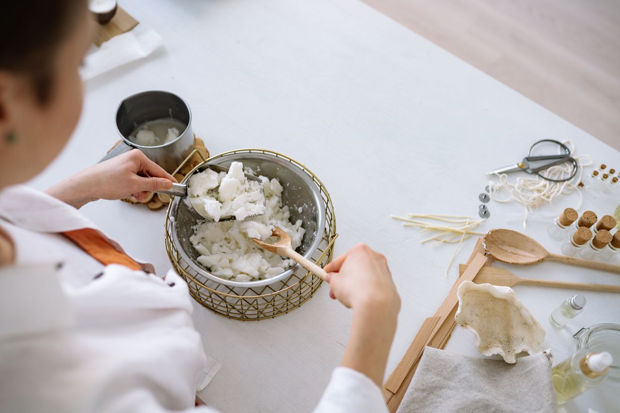 Person melting wax for candle making with tools on a white workspace.